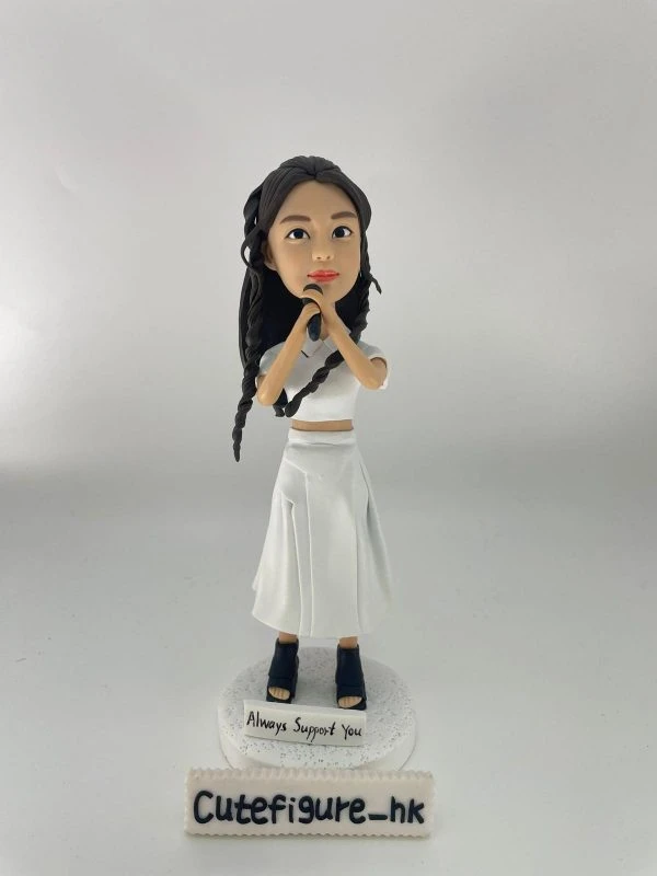 一款留有黑色長編髮的女歌手人像公仔，她身穿全白套裝，雙手握著咪高峰，站立在一個寫有 "Always Support You" 的白色圓形地台上。