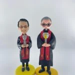 雙人牧師人像公仔：兩位人物身穿黑色長袍與紅色聖帶，領口白色牧領清晰；皆配戴眼鏡，微笑神情溫和。兩者手持銀色聖杯，聖帶上以金色與彩色圖案呈現麥穗、十字與象徵徽記。左側公仔站在圓形亮黃色底座，右側公仔站在略橢圓的黃色底座，整體質感細緻，適合作為教會場合致贈的禮物。