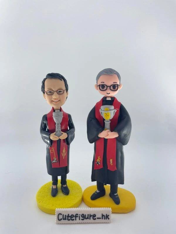 雙人牧師人像公仔：兩位人物身穿黑色長袍與紅色聖帶，領口白色牧領清晰；皆配戴眼鏡，微笑神情溫和。兩者手持銀色聖杯，聖帶上以金色與彩色圖案呈現麥穗、十字與象徵徽記。左側公仔站在圓形亮黃色底座，右側公仔站在略橢圓的黃色底座，整體質感細緻，適合作為教會場合致贈的禮物。
