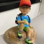 行山主題人像公仔：戴紅色帽子與眼鏡的男子公仔身穿藍色機能上衣、淺綠色短褲與灰色中筒襪，腳踩螢光綠黑色登山鞋，背負藍黑登山背包（側邊掛水瓶），坐在一塊帶有粗糙紋理的淺褐色岩石上休息；整體呈現輕裝郊遊與登山途中小憩的氛圍，適合熱愛行山者的客製人像公仔展示擺設。