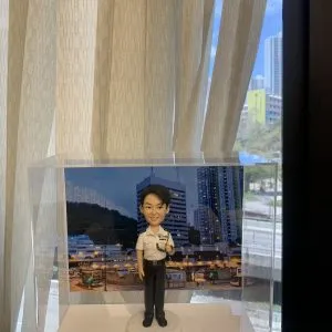 都會值勤意象融入典藏盒：本款警察人像公仔以白恤衫制服搭配通訊裝備為主視覺，臉部表情雕刻、髮絲刻線、肩章與口袋摺痕、對講機麥克風與線路、腰帶扣具等皆以手作成形並分色上色。圓形紋理底座配合城市夜景背板，凸顯專業而親和的巡邏風格。作為升遷誌慶、輪調餞別或部門致謝的送禮之選，兼具紀念與展示價值。