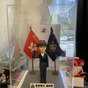 透明展示盒內的警察人像公仔：身著深藍制服與制帽，右手舉至眉間敬禮，左手自然下垂；胸前有肩帶與口袋釦飾，黑色皮鞋。人物站在灰色方形地台中央，左右各有一面旗幟：左側為紅底白花旗，右側為深藍色警徽旗。前方白底名牌印有中英對照的職級稱呼與姓名；名牌左側帶有警徽圖案。背板中央見到圓形紫色標誌，展示盒置於工作桌上，周圍有手工具與模型配件；窗外為高樓景觀。整體呈現正式、榮譽與頒贈性質，適合作為升遷、榮休或部門致意的送禮。