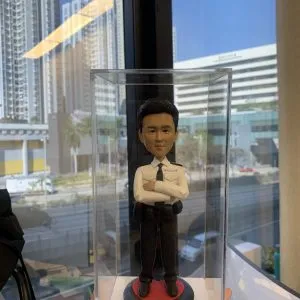 透明直立展示盒內放置一尊以「督察」職級為主題的警察人像公仔。公仔黑色短髮，神情沉穩；穿白色長袖制服襯衫，肩章與口袋翻蓋清晰，左胸可見「POLICE」字樣與名牌位置；配黑色領帶。腰間繫黑色多功能裝備腰帶，掛有對講機、套袋與工具袋；下身為深色長褲與黑色皮鞋。公仔雙臂於胸前交叉，站在黑紅相間的圓形台座上。台座前方有米金色名牌，印有英文姓名、祝賀升遷字樣與日期。展示盒由下方手掌托住，拍攝地點靠窗；窗外可見多棟高層住宅與辦公建築、道路與行道樹，藍天晴朗，玻璃上映出室內長條燈的反光，形成城市窗景與警務主題的對比。