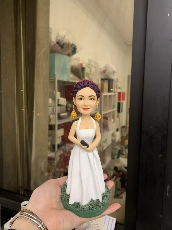 一款為擁有紫色編髮造型的女歌手訂製的人像公仔，她身穿白色長裙，配戴金色玫瑰耳環，手持咪高峰，並站立在一個綠色草地底座上。