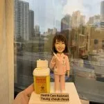 穿著粉紅色制服的醫護人像公仔，胸前掛有員工證與聽診器，手持注射器，旁邊放置一個黃色醫療廢物收集箱，上面貼有「DANGER」與生物危害標誌。底座標示「Health Care Assistant Yeung Cheuk Cheuk」。此公仔象徵專業醫護精神，適合作為感謝或紀念禮物。