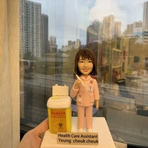 穿著粉紅色制服的醫護人像公仔，胸前掛有員工證與聽診器，手持注射器，旁邊放置一個黃色醫療廢物收集箱，上面貼有「DANGER」與生物危害標誌。底座標示「Health Care Assistant Yeung Cheuk Cheuk」。此公仔象徵專業醫護精神，適合作為感謝或紀念禮物。