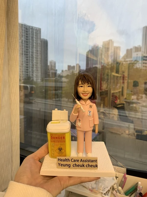 穿著粉紅色制服的醫護人像公仔，胸前掛有員工證與聽診器，手持注射器，旁邊放置一個黃色醫療廢物收集箱，上面貼有「DANGER」與生物危害標誌。底座標示「Health Care Assistant Yeung Cheuk Cheuk」。此公仔象徵專業醫護精神，適合作為感謝或紀念禮物。