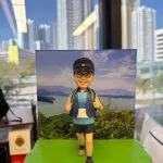 越野跑手人像公仔，戴著黑色帽子與眼鏡，身穿運動背心、短褲與登山鞋，背著背包踏步向前。背景是山脈與湖泊風景，展現戶外越野長跑的熱情與活力。