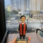 單人牧師人像公仔：黑色長袍搭配紅色聖帶，內襯白色立領，短黑髮、微笑表情端莊親切；站立於灰色石板質感底座，上面兩側為綠色草地與小花點綴，前方附銘牌可刻姓名與年份（範例顯示「牧師榮休 2025」字樣）。整體細節清晰，適合作為榮休、授職與致謝的人像公仔禮物。