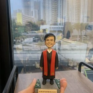 單人牧師人像公仔：黑色長袍搭配紅色聖帶，內襯白色立領，短黑髮、微笑表情端莊親切；站立於灰色石板質感底座，上面兩側為綠色草地與小花點綴，前方附銘牌可刻姓名與年份（範例顯示「牧師榮休 2025」字樣）。整體細節清晰，適合作為榮休、授職與致謝的人像公仔禮物。