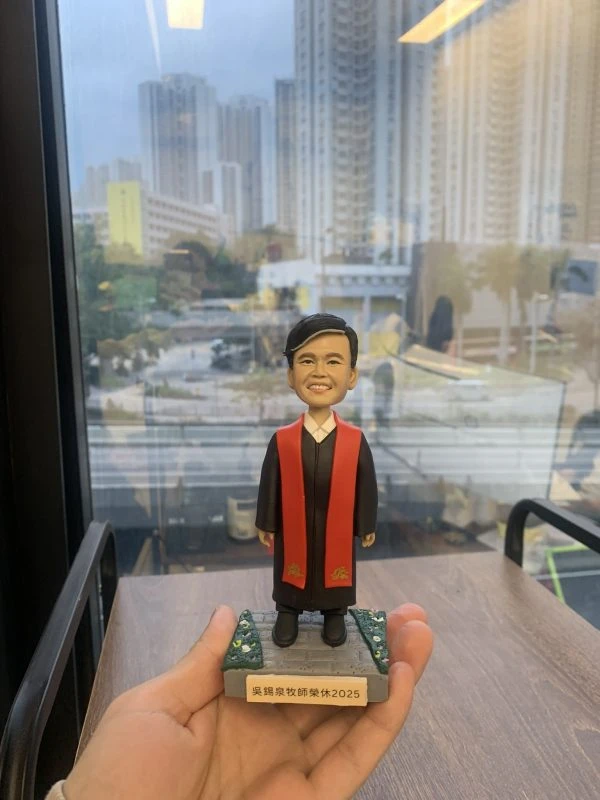 單人牧師人像公仔：黑色長袍搭配紅色聖帶，內襯白色立領，短黑髮、微笑表情端莊親切；站立於灰色石板質感底座，上面兩側為綠色草地與小花點綴，前方附銘牌可刻姓名與年份（範例顯示「牧師榮休 2025」字樣）。整體細節清晰，適合作為榮休、授職與致謝的人像公仔禮物。