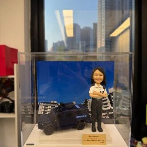 透明壓克力展示盒中,立有一尊女性「督察」主題的警察人像公仔。公仔短黑髮、微笑神情,雙臂在胸前交叉;上身穿白色短袖制服襯衫,肩章與名牌位置清晰,左胸別有對講機肩咪與連線;下身為黑色戰術長褲與黑色長靴,腰間繫有裝備腰帶。人物站於淺色紋理底座,前方設有金色名牌,印有祝賀與致謝文字。背板是一輛深藍色多用途裝甲車(POLICE標示清楚)與幾何線條感強烈的建築群;盒外環境靠近窗邊,窗外可見密集住宅高樓與藍天,室內長條燈在玻璃上留有反光。整體營造出戰術勤務與城市場景結合的紀念展示與送禮氛圍。
