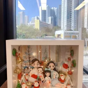 手工製作的 人像公仔 場景作品,裝在透明展示盒中,呈現一家四口的溫馨畫面。父母與兩個小孩坐在一起,背景裝飾有聖誕樹、金色小熊、禮物盒及多個聖誕老人公仔,搭配燈飾與紅色毛帽小物,營造節日氛圍。此作品結合 家庭團聚 與 聖誕節禮物 主題,展現專屬紀念與溫馨心意。