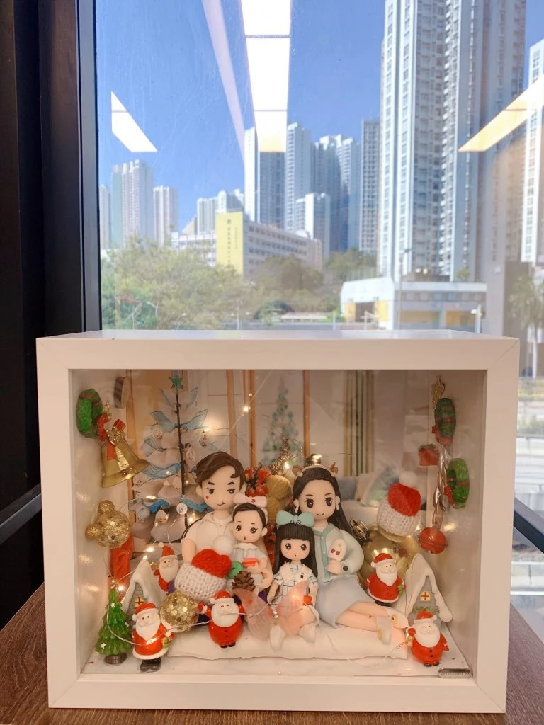 手工製作的 人像公仔 場景作品，裝在透明展示盒中，呈現一家四口的溫馨畫面。父母與兩個小孩坐在一起，背景裝飾有聖誕樹、金色小熊、禮物盒及多個聖誕老人公仔，搭配燈飾與紅色毛帽小物，營造節日氛圍。此作品結合 家庭團聚 與 聖誕節禮物 主題，展現專屬紀念與溫馨心意。