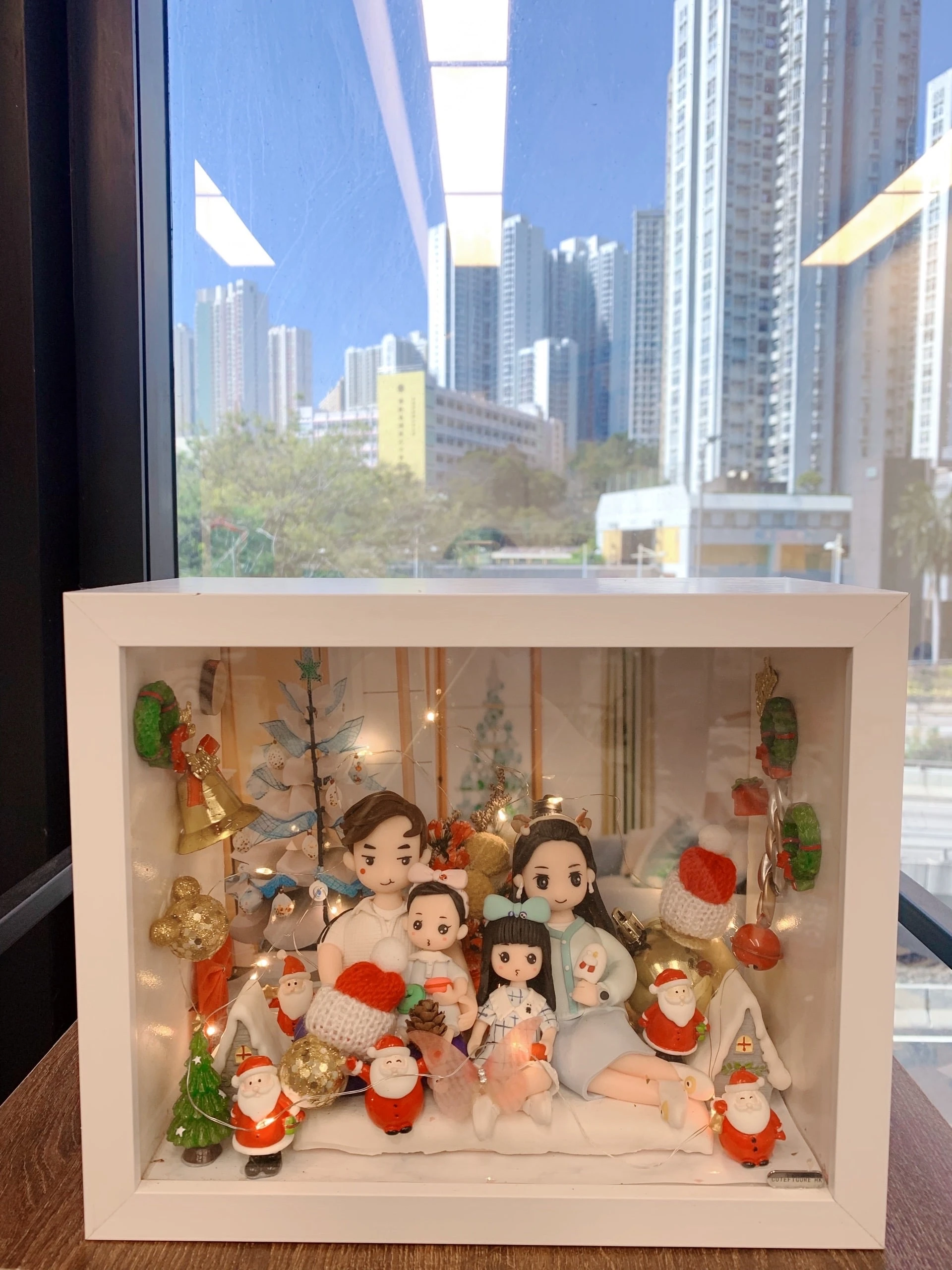 手工製作的 人像公仔 場景作品，裝在透明展示盒中，呈現一家四口的溫馨畫面。父母與兩個小孩坐在一起，背景裝飾有聖誕樹、金色小熊、禮物盒及多個聖誕老人公仔，搭配燈飾與紅色毛帽小物，營造節日氛圍。此作品結合 家庭團聚 與 聖誕節禮物 主題，展現專屬紀念與溫馨心意。