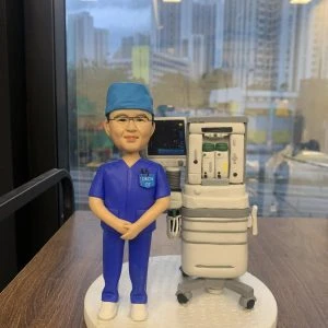 訂製人像公仔：一位身穿深藍色手術服與手術帽的醫生，佩戴眼鏡，站立於麻醉機旁邊，機器上附有顯示監測螢幕與氣體供應裝置，重現手術室真實臨床場景，展現醫護專業。