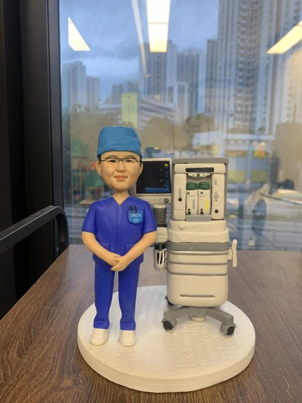 訂製人像公仔：一位身穿深藍色手術服與手術帽的醫生，佩戴眼鏡，站立於麻醉機旁邊，機器上附有顯示監測螢幕與氣體供應裝置，重現手術室真實臨床場景，展現醫護專業。