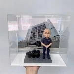 這件作品以立正合掌站姿的警察人像公仔為主角，頭部比例略大以突顯親切神情，臉型、眉眼與耳部轉折以手作雕刻呈現自然起伏，禿頂髮際、眼鏡框與唇線描繪細膩；深藍制服的口袋、肩章與縫線以細筆補色，褲管折與鞋面高光加強了質感。地台採六角石紋塑形，左側配置黑色警用裝甲車模型，含「POLICE」字樣與車頂艙口等硬派元素，與背板的政府大樓與旗幟相呼應，形成完整的警務情境。透明展示盒有效防塵並提升陳列儀式感，適合作為榮升、退休、嘉許或周年紀念的體面之選，亦非常適合同事與親友送禮；可進一步客製姓名、職級、頒贈日期與隊章背板，或增添對講機、勳表、證件夾等配件，完整展現警察人像公仔的專業與情感價值。