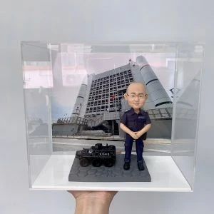 這件作品以立正合掌站姿的警察人像公仔為主角,頭部比例略大以突顯親切神情,臉型、眉眼與耳部轉折以手作雕刻呈現自然起伏,禿頂髮際、眼鏡框與唇線描繪細膩;深藍制服的口袋、肩章與縫線以細筆補色,褲管折與鞋面高光加強了質感。地台採六角石紋塑形,左側配置黑色警用裝甲車模型,含「POLICE」字樣與車頂艙口等硬派元素,與背板的政府大樓與旗幟相呼應,形成完整的警務情境。透明展示盒有效防塵並提升陳列儀式感,適合作為榮升、退休、嘉許或周年紀念的體面之選,亦非常適合同事與親友送禮;可進一步客製姓名、職級、頒贈日期與隊章背板,或增添對講機、勳表、證件夾等配件,完整展現警察人像公仔的專業與情感價值。