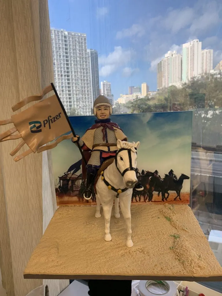 英勇騎士主題人像公仔，身穿盔甲與斗篷的男性公仔騎乘白馬，手持印有 Pfizer 標誌的旗幟，背景為沙場戰馬場景。整體設計充滿英勇與榮耀氛圍，專為老闆退休製作的獨特禮物。