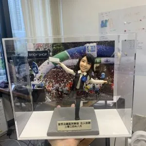 以手作定格高光時刻：這款警察人像公仔採用動態展臂站姿與單腳微踮設計，臉部眉眼弧度、嘴角微笑、髮絲刻線和背心/襯衫的摺痕都逐一刻畫。背板選用大型活動現場影像，與透明展示盒共同營造儀式感；灰色質感底座與金色牌匾則強化典藏層次。無論作為升遷誌慶、團隊紀念，或致謝同事的專屬送禮，皆能體現專屬故事與心意。