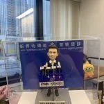 精緻手工製作的警務人員人像公仔（人像公仔、警察人像公仔），置於透明亞加力展示盒內；公仔五官雕刻細膩，穿白色恤衫、黑色戰術背心、黑褲與黑鞋，站在灰色石紋底座上。前方設多支新聞咪高峰連不同媒體咪牌，咪線以紅、藍、黑線纜呈現；中央有編號牌「NTNRN 22000502」。底座金屬牌刻字「新界北總區刑事部 友人送贈 二零二四年八月」。背板印有「新界北總區 警察總部 NEW TERRITORIES NORTH REGIONAL HEADQUARTERS」字樣；背景為室內辦公環境與窗外高樓，右側可見毛絨公仔擺設。整體做工精美、塗裝乾淨，還原警方記者會發言場景。