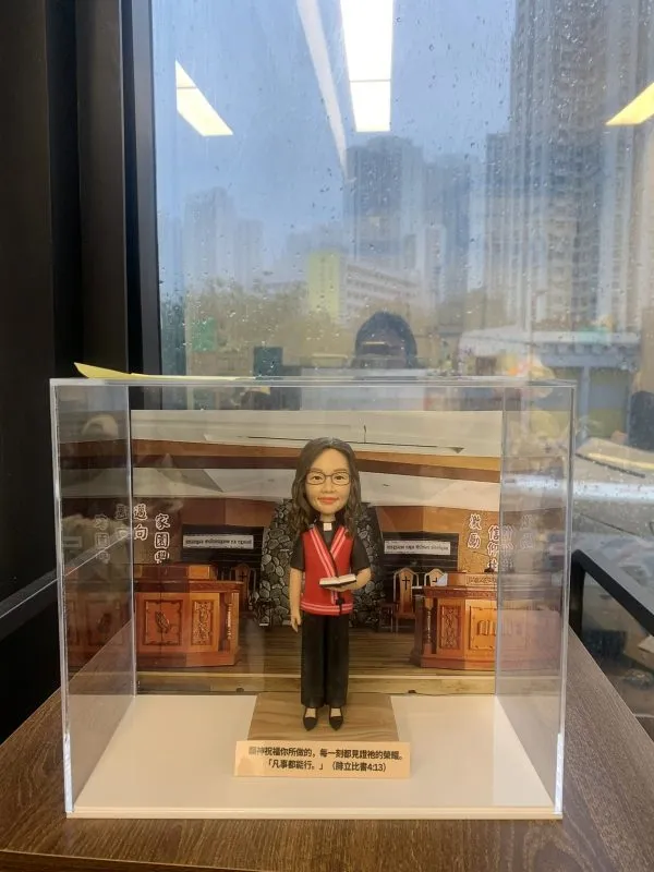 單人牧師人像公仔置於透明展示盒中：角色戴眼鏡、微笑神情，穿黑色服飾搭配紅色聖帶與白色牧領，手持書本；背景為教堂講台與木質裝飾的場景板，底座前方附有金色銘牌可刻經文或祝福語。整體精緻，適合作為教會授職、周年、榮休與致謝的禮物。