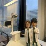 兩位身穿白袍的醫生人像公仔，站立於醫療場景模型前，背景包含電腦顯示屏與手術燈。公仔細節精緻，展現專業醫護形象，腳踩藍色鞋套，象徵無菌醫療環境。底座刻有「于春紅醫生」字樣，這款專屬定製醫護公仔極具收藏與送禮價值。