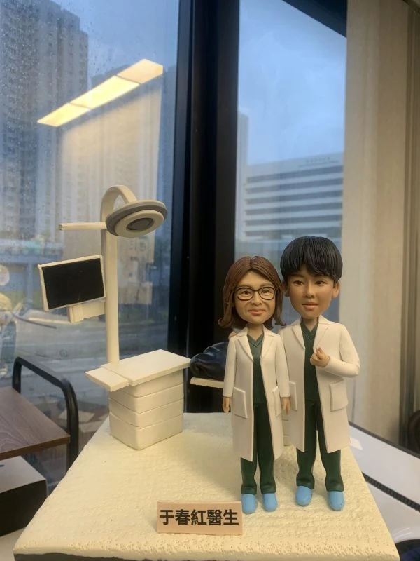 兩位身穿白袍的醫生人像公仔，站立於醫療場景模型前，背景包含電腦顯示屏與手術燈。公仔細節精緻，展現專業醫護形象，腳踩藍色鞋套，象徵無菌醫療環境。底座刻有「于春紅醫生」字樣，這款專屬定製醫護公仔極具收藏與送禮價值。