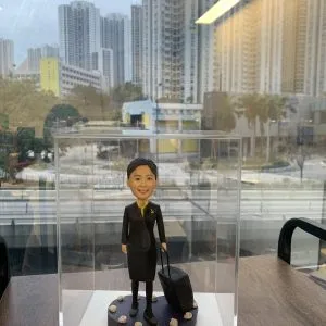 一款為名為Honey的航空空姐訂製的2025年榮休人像公仔，公仔身穿制服並拖著一個行李箱作為背景配件裝飾，放置於透明展示盒中。