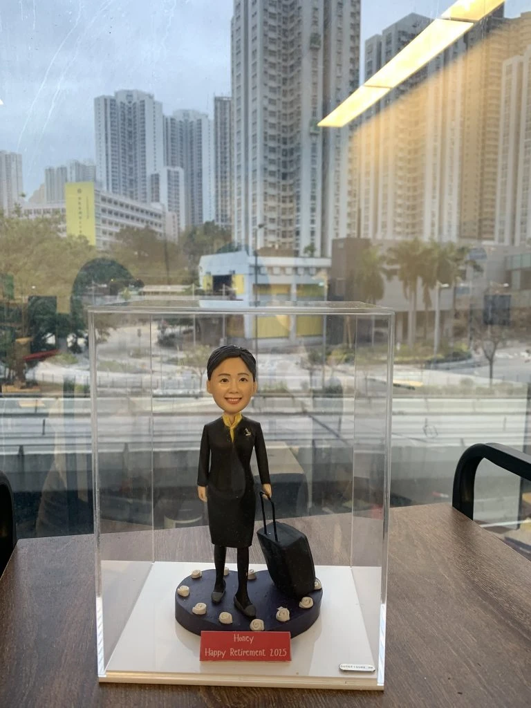 一款為名為Honey的航空空姐訂製的2025年榮休人像公仔，公仔身穿制服並拖著一個行李箱作為背景配件裝飾，放置於透明展示盒中。