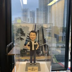 透明直立展示盒中擺放一尊便衣風格的「偵緝警員」主題人像公仔。人物黑色短髮、微笑表情,雙手插腰站姿輕鬆自信;上身穿白色短袖POLO衫,外搭黑色無袖戰術背心,左胸貼有「POLICE」與名牌識別貼;下身為深色長褲與黑色便鞋。腳下是灰色六角石鋪面造型台座,前方金色名牌以繁體中文寫有姓名與「榮休之樂 2025」等致意文字。背板為十字路口前的高樓商業區外牆與玻璃帷幕街景,地面見黃格線路口與行人道;展示盒由下方手掌托起,外部環境靠窗,窗外可見高密度住宅大廈與道路車流,窗玻璃映出室內長條燈反光與書架物件。