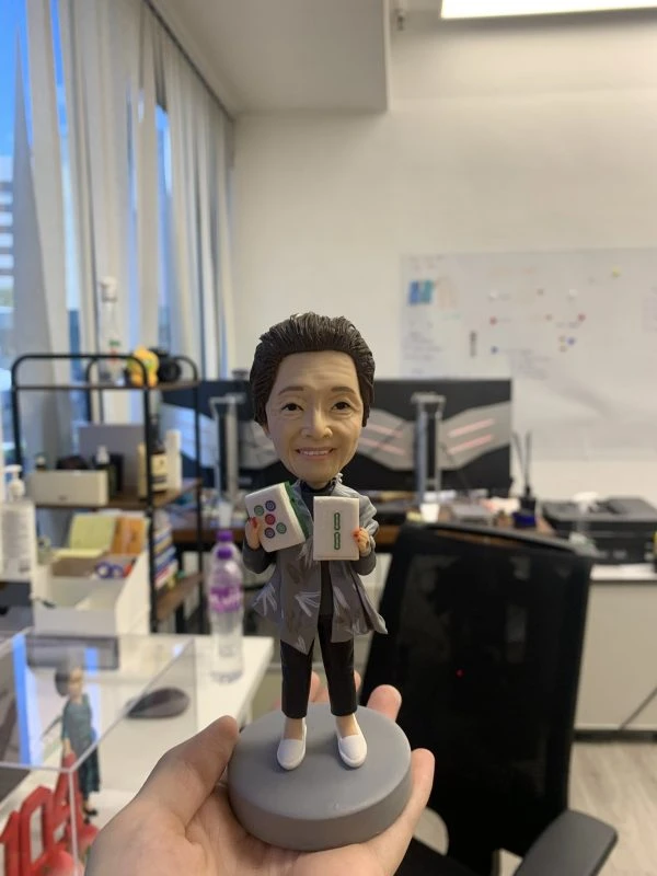 手工訂製的人像公仔，以長者形象為原型雕刻，手持兩張大型麻將牌「八筒」與「八萬」，展現真實細節與趣味設計。此人像公仔將麻將文化融入專屬個人化收藏，不僅象徵吉祥好運，也成為麻將愛好者與長輩專屬的獨特禮物。