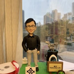 一個男同事的人像公仔站立在高爾夫主題造景模型上，身穿黑色襯衫與灰色長褲，旁邊有高爾夫球袋、球具與標語牌，牌子上寫著「華仔 Enjoy your Life」。