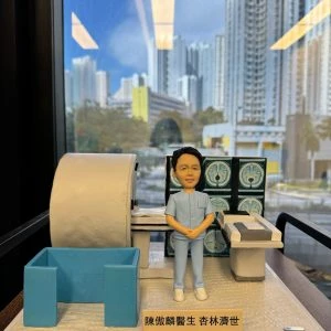 透明展示盒內的人像公仔：一位身穿淺藍色醫療制服的男醫生雙手合於腹前，身旁擺放著一台大型醫療影像檢查設備（MRI 磁力共振掃描機），後方螢幕顯示多張腦部斷層掃描影像，整體場景呈現放射科醫生的臨床專業環境。
