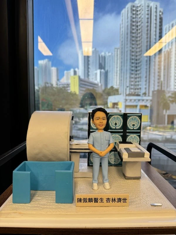 透明展示盒內的人像公仔：一位身穿淺藍色醫療制服的男醫生雙手合於腹前，身旁擺放著一台大型醫療影像檢查設備（MRI 磁力共振掃描機），後方螢幕顯示多張腦部斷層掃描影像，整體場景呈現放射科醫生的臨床專業環境。