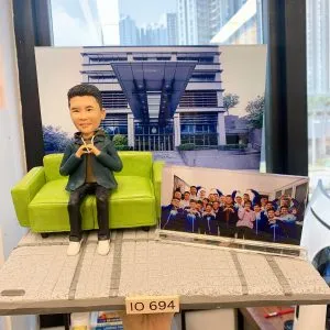 一款以警察為主題的人像公仔，身穿便服坐在綠色沙發上，身旁展示著與同袍的合照，背景是警察相關建築，底座前方印有「IO 694」字牌，象徵個人專屬身份與紀念。