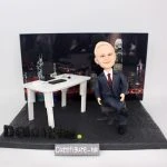 一款為外國老闆訂製的退休禮物人像公仔，公仔是一位穿西裝的男士，坐在辦公室的立體場景中，背景是香港維港背景夜景，底座上有Deloitte公司標誌。