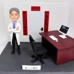 訂製醫生人像公仔，角色穿著白色醫生袍，雙手交叉站在辦公桌旁，桌上有電腦螢幕、鍵盤與滑鼠，背景寫有醫療對聯「剪扎切縫除病災、開膛訂腹癒百病」。底座標示「博愛醫院 Pok Oi Hospital」，象徵醫生的專業與醫療貢獻，適合作為榮休、紀念或醫護禮物。