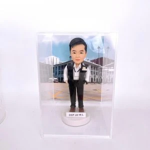 置於透明展示盒中的警察人像公仔：男性造型短髮，身著白色襯衫、深色長褲與黑色戰術背心，胸前別有「POLICE」識別章與名牌，雙手插袋站姿挺直；腳穿棕色鞋，立於白色圓形底座，前方貼有姓名縮寫標籤「DSIP LAI W.L.」。背景為戶外部門建築與廣場圖像，地面有黃色標線與旗桿，營造執勤場景氛圍。整體在純白環境中拍攝，突顯公仔細節與升職紀念意義。