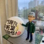 穿手術帽與深藍手術服的麻醉科醫生主題人像公仔，外搭綠色外套，右手指向一個寫有「屯門醫院 手術室 歡迎您！」的大型白色球形展示物；底座為灰色磚路與綠色草地點綴，背景是窗外高樓天際線。適合作為醫護致謝或退休紀念禮物。