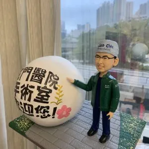 穿手術帽與深藍手術服的麻醉科醫生主題人像公仔,外搭綠色外套,右手指向一個寫有「屯門醫院 手術室 歡迎您!」的大型白色球形展示物;底座為灰色磚路與綠色草地點綴,背景是窗外高樓天際線。適合作為醫護致謝或退休紀念禮物。