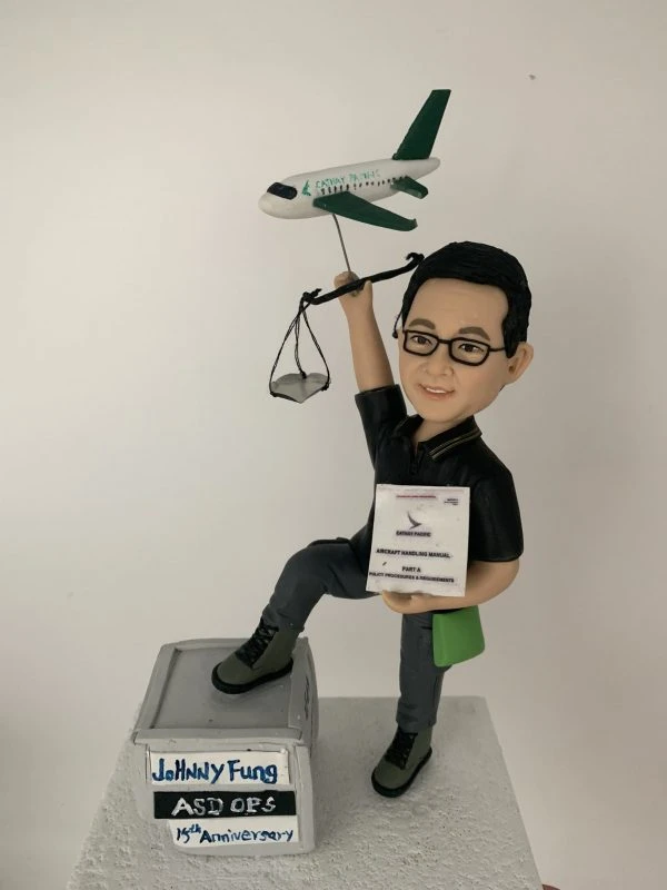 一款為國泰航空地勤營運人員訂製的15週年紀念人像公仔，主角手持飛機裝卸手冊，並舉著一個帶有飛機模型與天秤的背景配件裝飾。