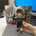 一款為Tiger Sir訂製的退休人像公仔，公仔身穿印有「Tiger」字樣的單車服，做出衝線勝利手勢，背景是消防局照片，底座銘牌寫有「Happy Retirement to Tiger Sir 2024」。