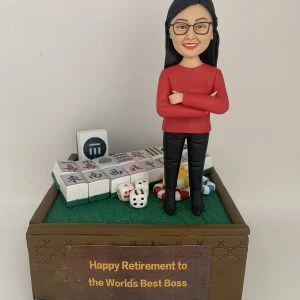 專屬訂製的人像公仔,以女性上司為原型,身穿紅色上衣與黑色長褲,雙手交叉微笑站立。底座設計成麻將桌風格,擺放多張麻將牌、骰子與籌碼,前方木製牌匾寫有「Happy Retirement to the World's Best Boss」,象徵榮退賀禮與專屬紀念。此人像公仔將 麻將元素 與感謝心意融合,打造獨特的退休祝福禮物。