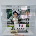 訂製醫護人像公仔，角色身穿白色醫護制服，佩戴名牌，手持聽診器，背景為九龍醫院大樓，底座印有「Kowloon Hospital」標誌。前方刻有「Happy Retirement Dr Rosanna Chau, Department Manager (PT), Kowloon Hospital」，象徵醫護人員榮休紀念。