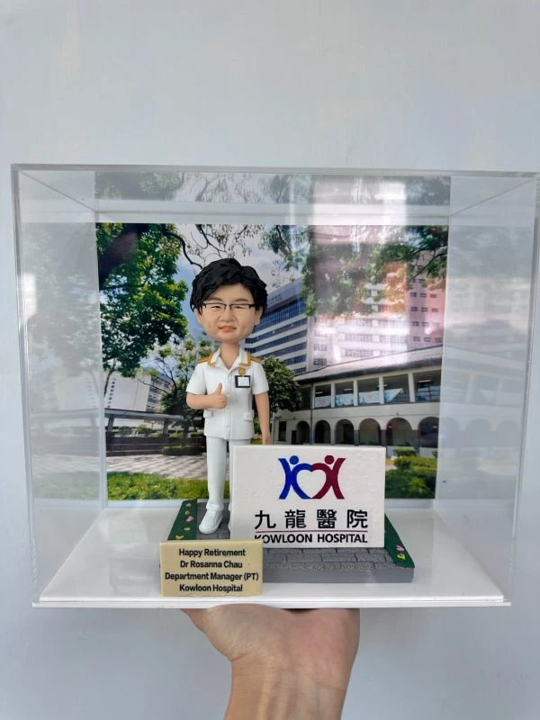 訂製醫護人像公仔，角色身穿白色醫護制服，佩戴名牌，手持聽診器，背景為九龍醫院大樓，底座印有「Kowloon Hospital」標誌。前方刻有「Happy Retirement Dr Rosanna Chau, Department Manager (PT), Kowloon Hospital」，象徵醫護人員榮休紀念。