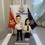 一款祝賀警務處行動處處長晉升的雙人版人像公仔。男公仔身穿警官制服，女公仔身穿便服，站立在印有警務處標誌及職銜的背景板前，兩旁分別是香港區旗和警隊旗幟。底座銘牌刻有祝賀晉升的字句。