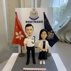 一款祝賀警務處行動處處長晉升的雙人版人像公仔。男公仔身穿警官制服，女公仔身穿便服，站立在印有警務處標誌及職銜的背景板前，兩旁分別是香港區旗和警隊旗幟。底座銘牌刻有祝賀晉升的字句。