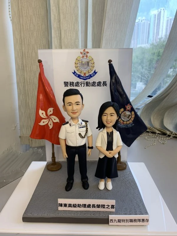 一款祝賀警務處行動處處長晉升的雙人版人像公仔。男公仔身穿警官制服，女公仔身穿便服，站立在印有警務處標誌及職銜的背景板前，兩旁分別是香港區旗和警隊旗幟。底座銘牌刻有祝賀晉升的字句。